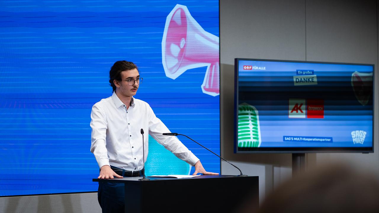 Es wird ein junger Mann im weißen Hemd beim Halten seiner Rede gezeigt. 